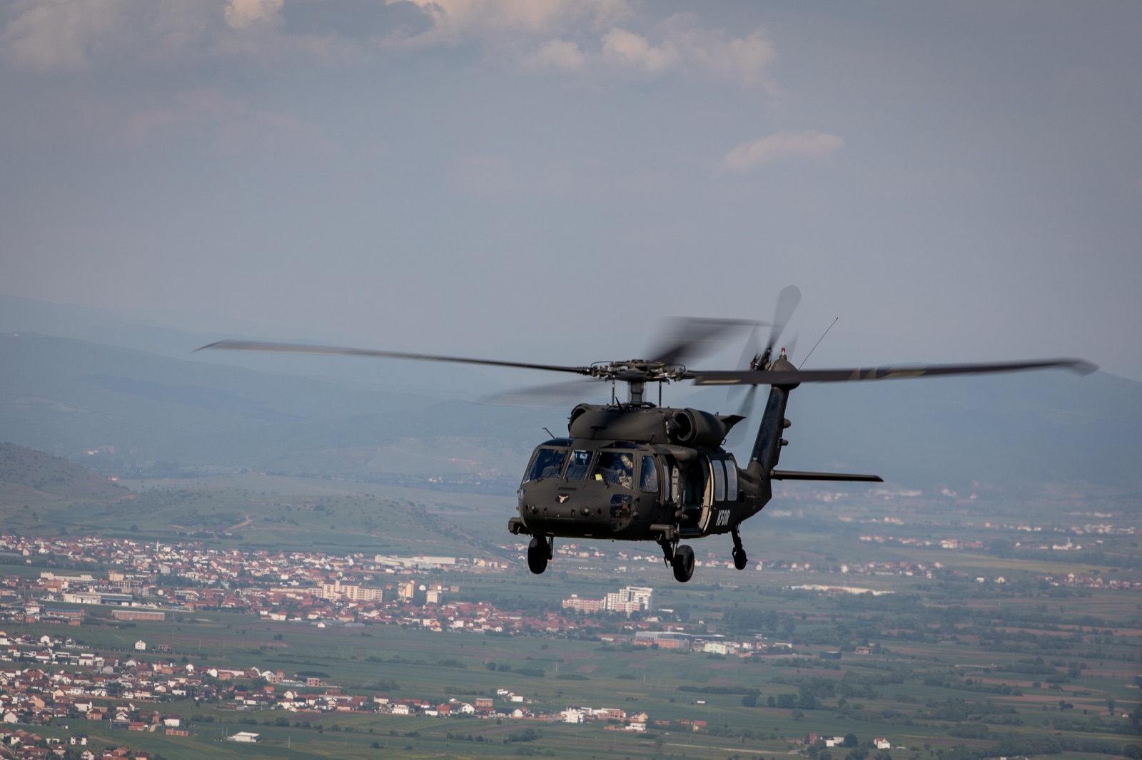A dark green UH-60 Black Hawk helicopter in flight above a town in a wide valley