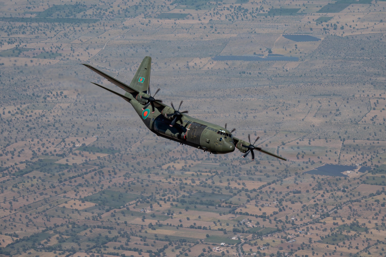 A green Bangladesh Air Force C-130J Hercules banking over a chequerboard of fields