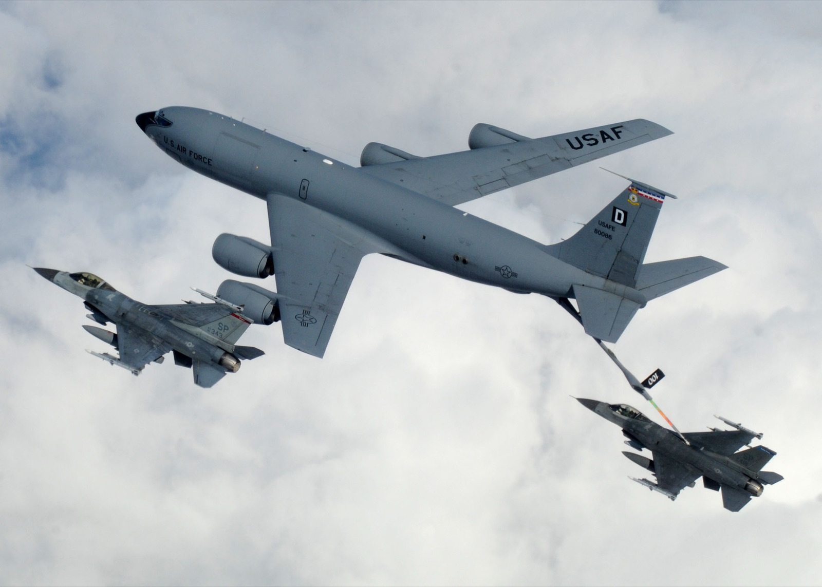 A grey KC-135 tanker with its boom lowered into an F-16 flying beneath its tail, a second F-16 alongside