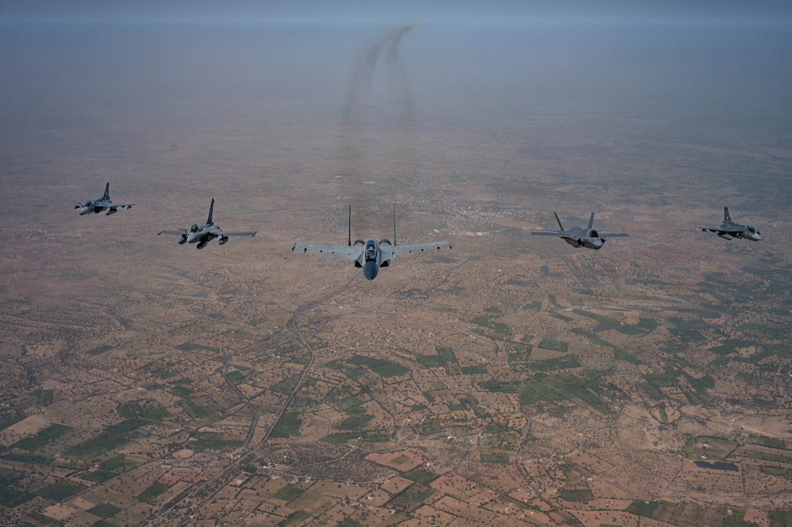 Five fighters in line abreast over a dusty plain: two Tejas, a French Rafale, an Indian Su-30MKI and an American F-35A