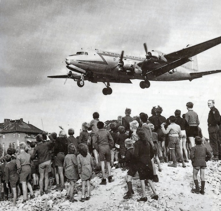 Children and a few adults stand on a mound of rubble and watch a four-engined C-54 transport pass low overhead with its wheels down