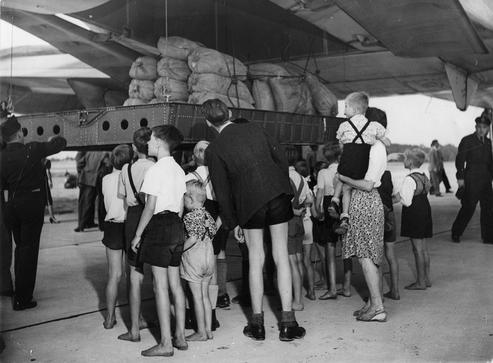 A crowd of children, many barefoot and in shorts, stand under the wing of a huge aircraft looking up at a steel cargo platform stacked with sacks of flour