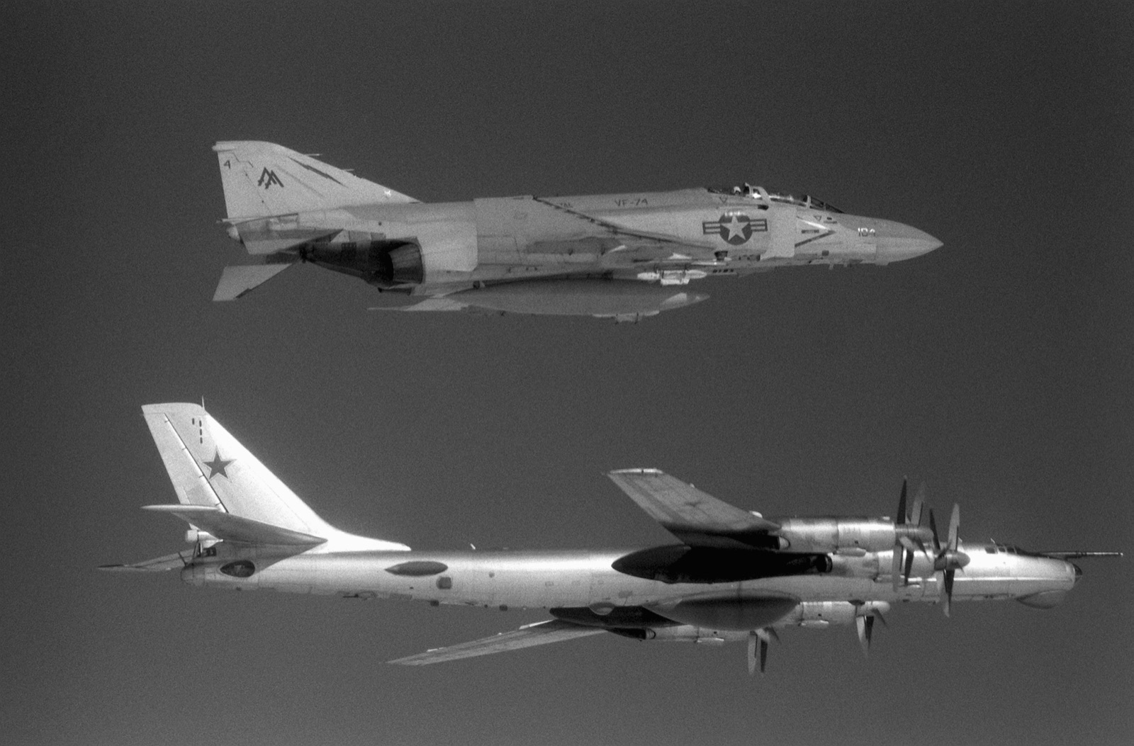 A grey US Navy F-4 Phantom flies in formation above a much larger Soviet Tu-95 Bear bomber with swept wings and contra-rotating propellers