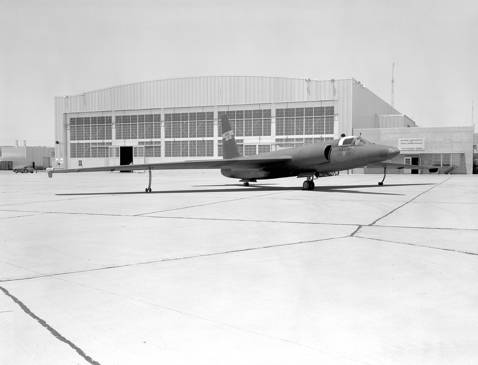A dark, long-winged U-2 stands on a concrete apron in front of a large hangar, its tail marked NASA and the serial 55741