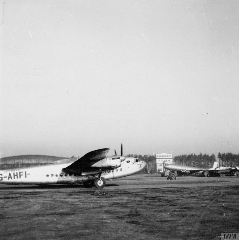 A four-engined Avro York airliner registered G-AHFI taxis across a grass airfield, with a control tower and other transport aircraft behind
