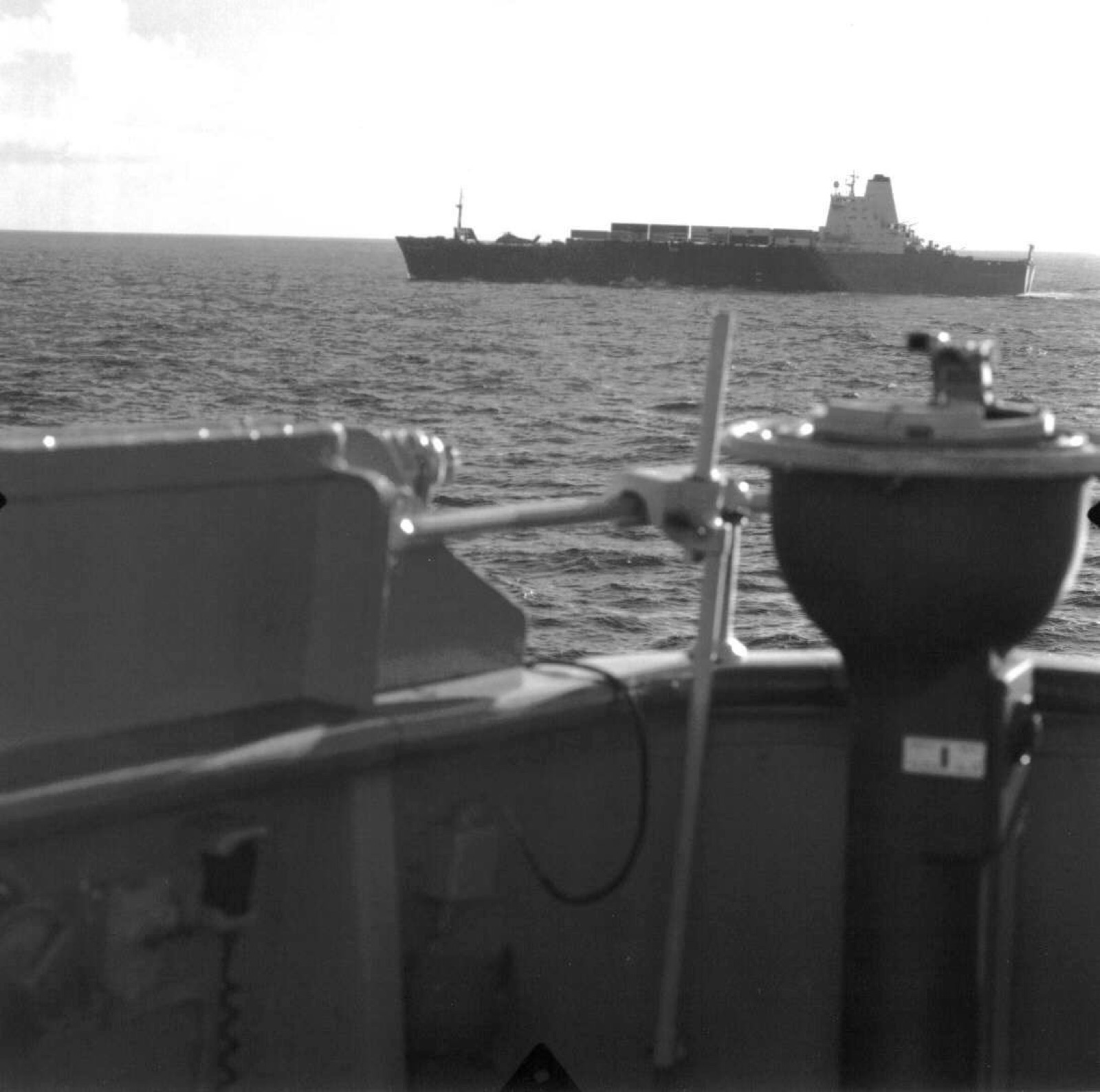 A long dark container ship on the horizon, seen past the out-of-focus fittings of a warship’s bridge wing