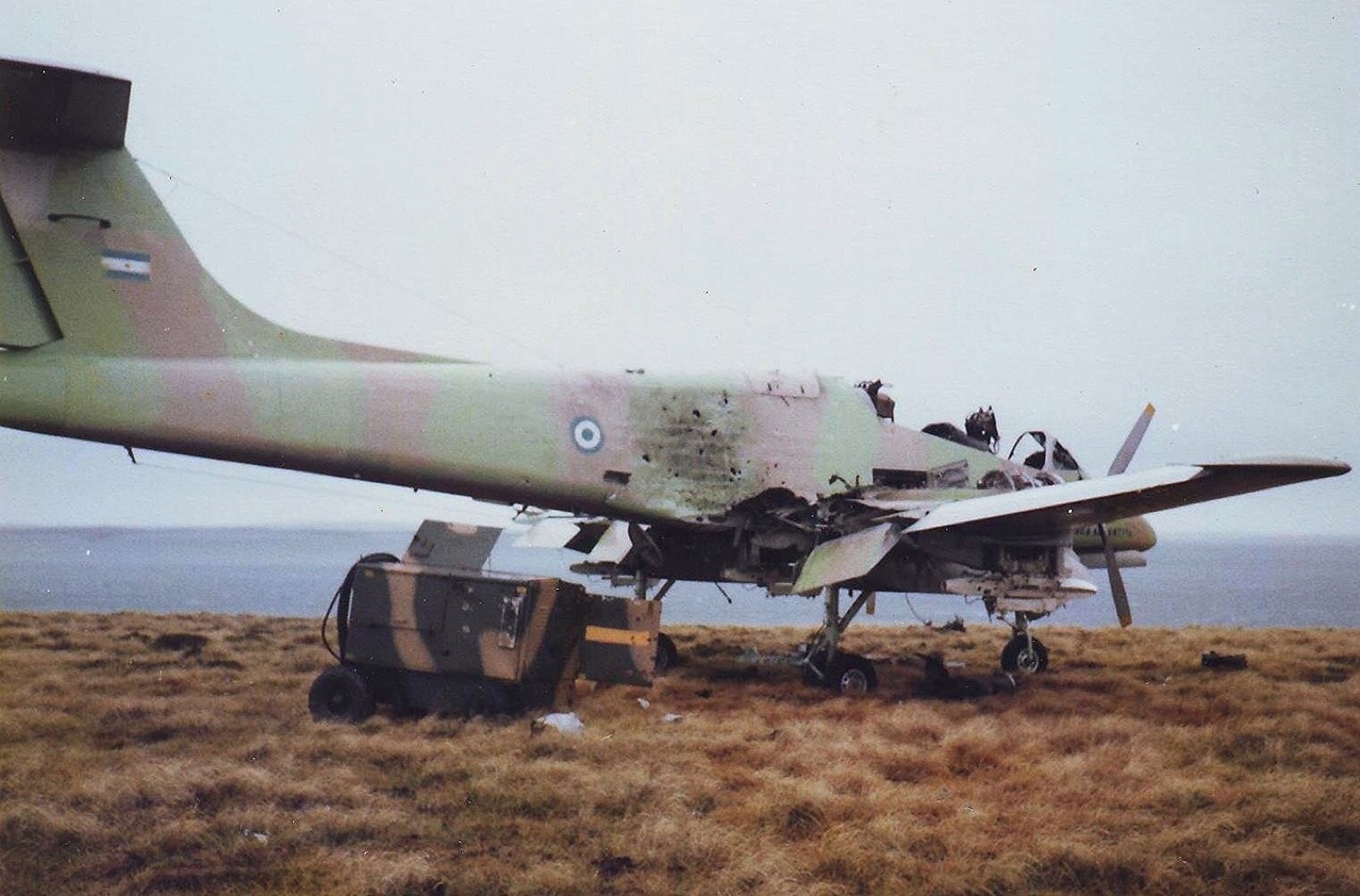 A wrecked camouflaged twin-engined Pucará on grass by the sea, its nose and engine torn open, a ground-equipment trolley beside it