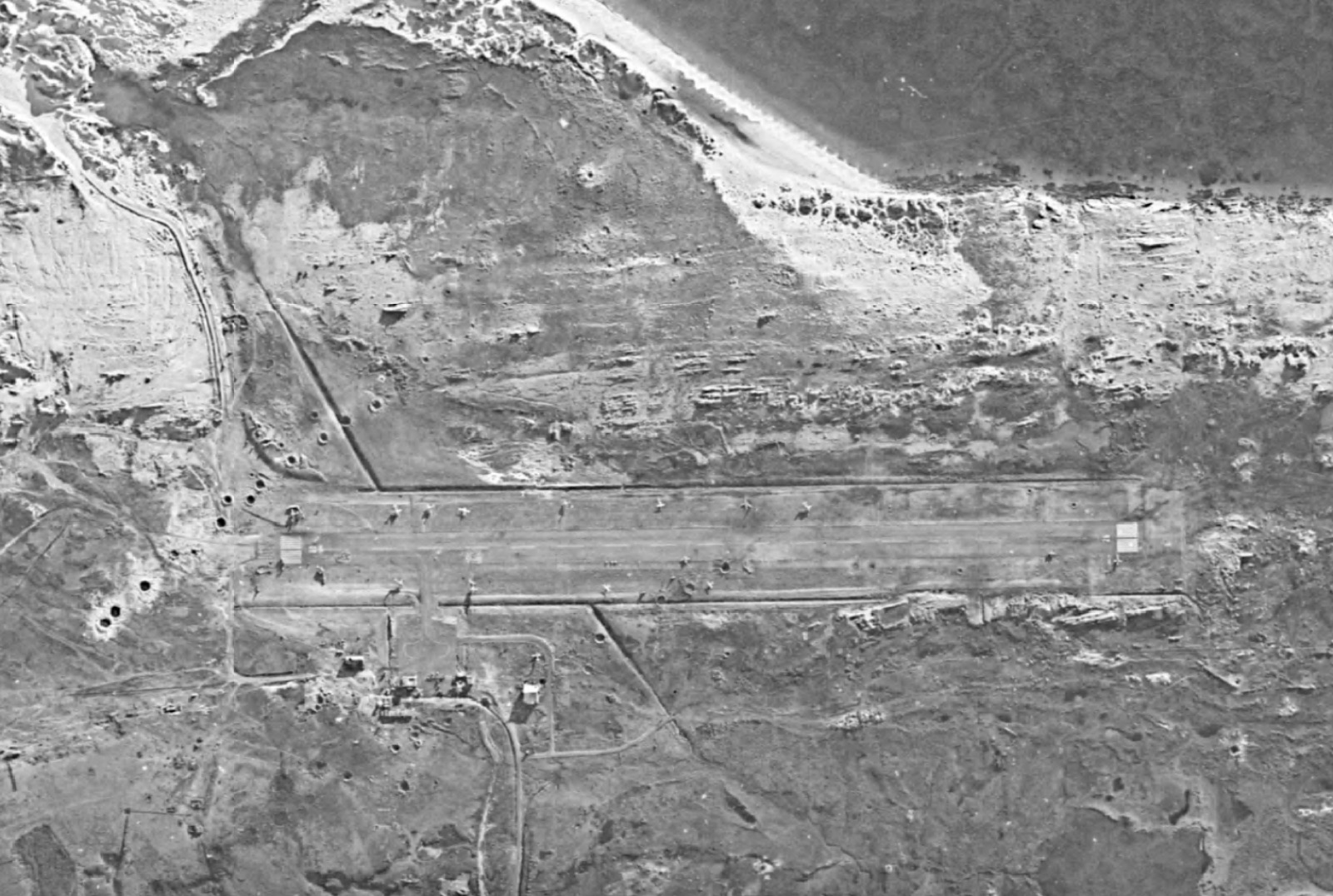 Black-and-white satellite view of Stanley airfield: a single runway running left to right, pocked with craters at its western end