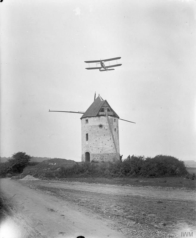 A B.E.2c biplane flies low over a stone windmill with broken sails beside a country road