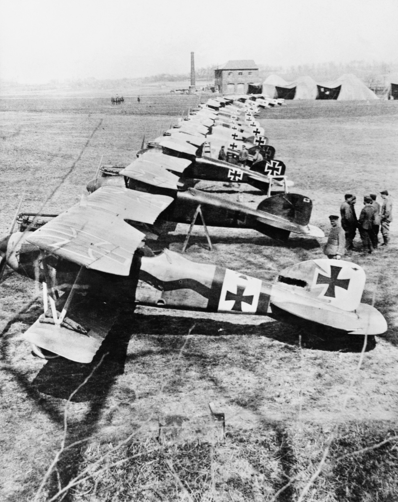A long diagonal line of German Albatros D.III biplane fighters parked on a grass airfield, crosses on their fuselages and tails, with tent hangars and a brick building beyond
