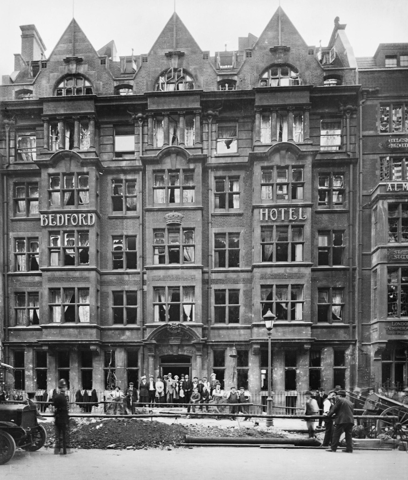 The front of the Bedford Hotel, a tall Victorian brick building with most of its windows blown out; guests stand on the steps while workmen dig at a crater in the road