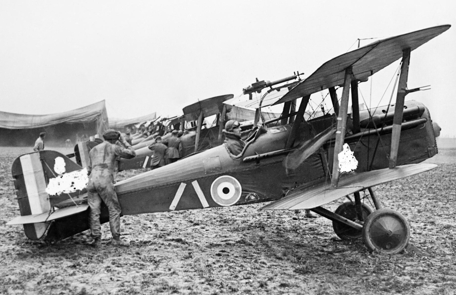 A row of S.E.5a fighters on a muddy airfield, the nearest with its pilot in the cockpit and a mechanic in overalls at the tail; the serial number has been scratched out of the negative