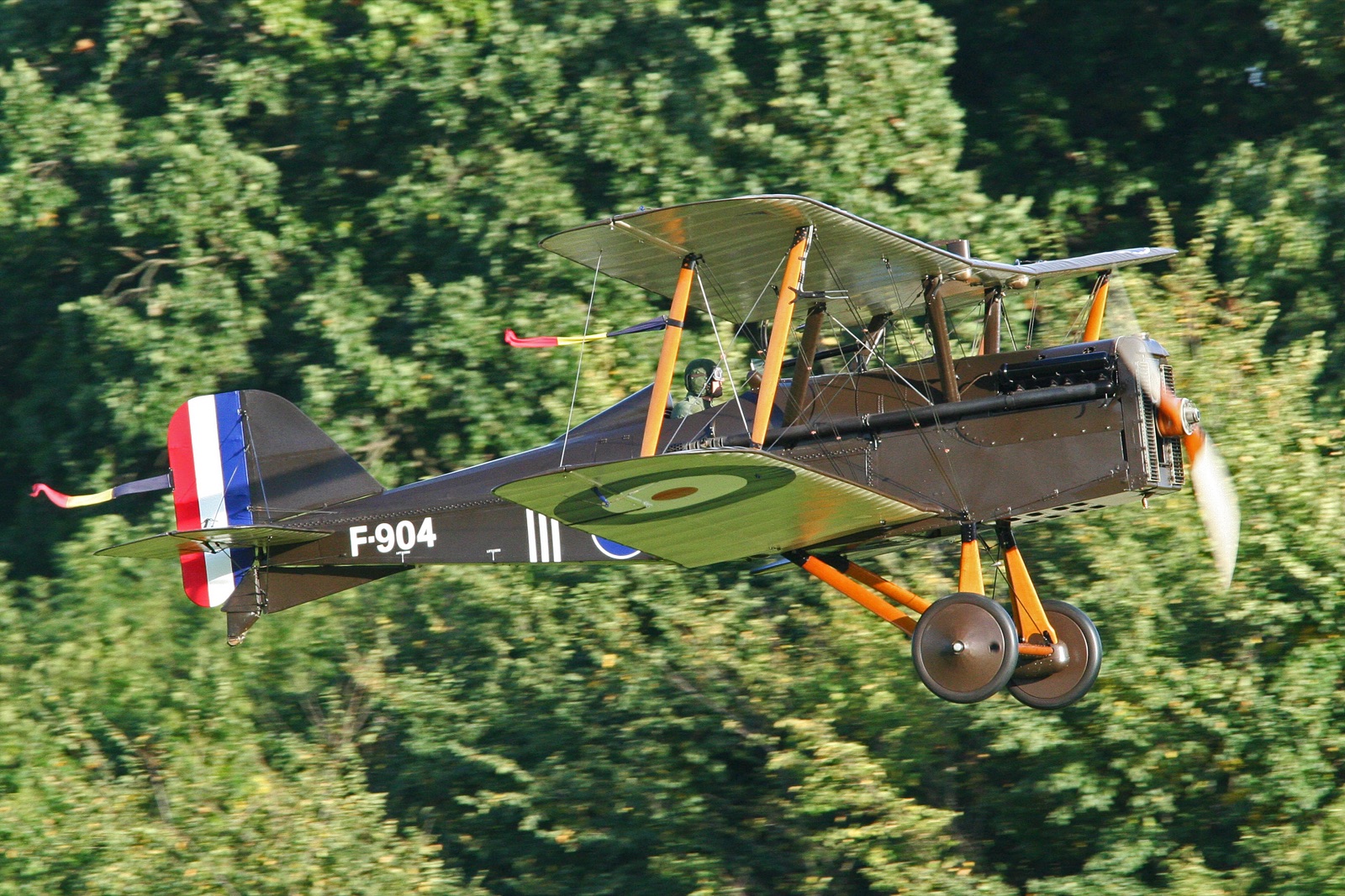A brown-painted S.E.5a biplane with red, white and blue tail stripes and the serial F-904 flies low past a backdrop of trees