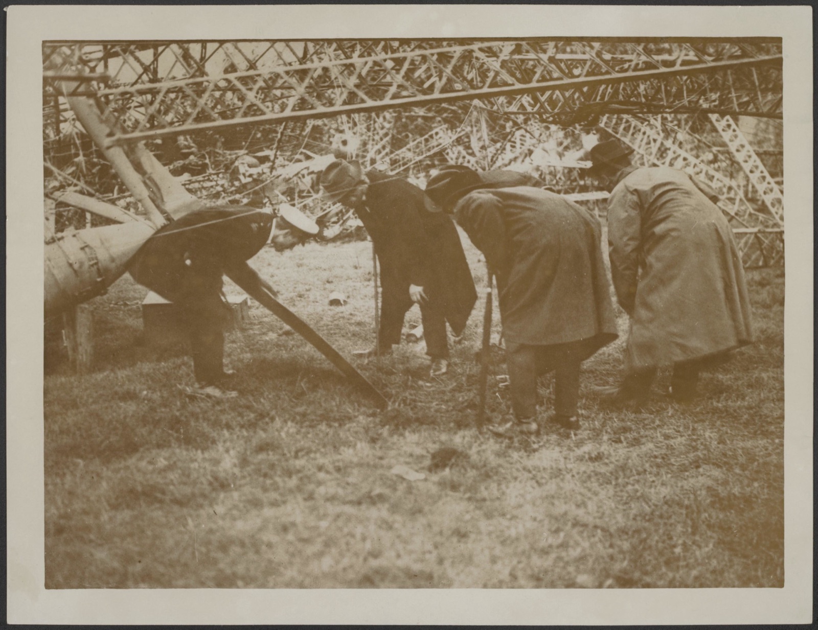 Four men in overcoats and hats stoop to look at the ground beneath the tangled lattice girders of a wrecked airship