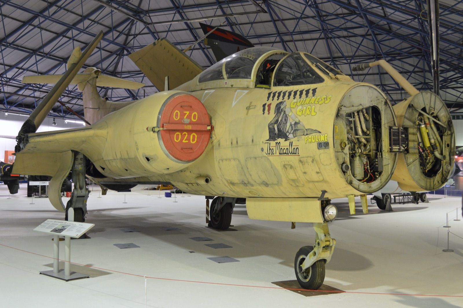 A desert-pink Buccaneer in a museum hangar, nose radome swung open, with bomb symbols and nose art under the cockpit