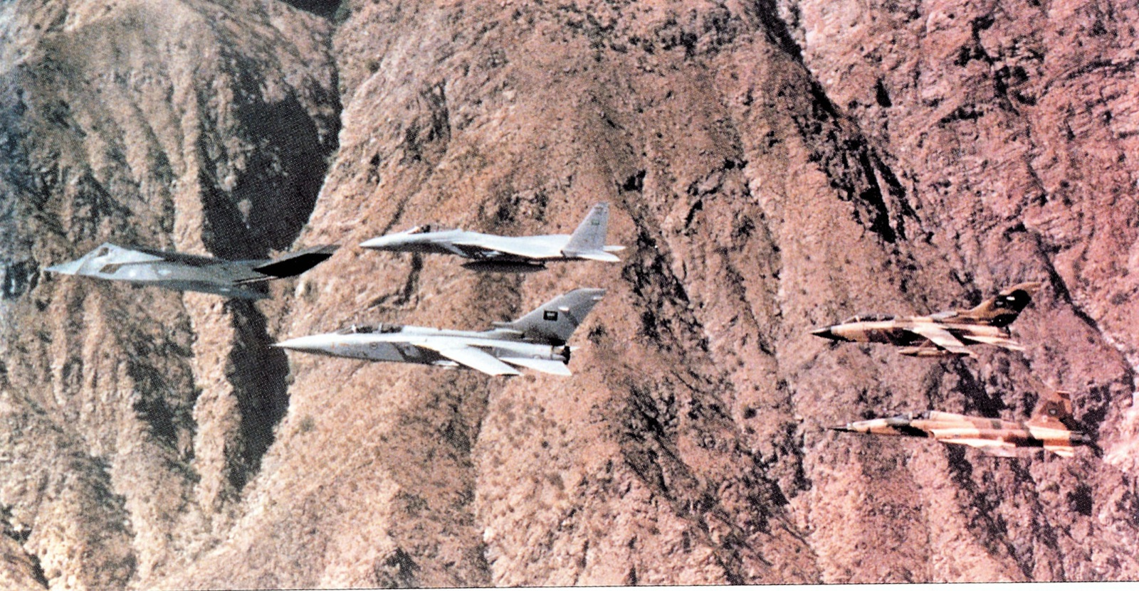 A black faceted F-117A leads a formation of Saudi F-15, Tornado and two F-5 fighters over red mountains