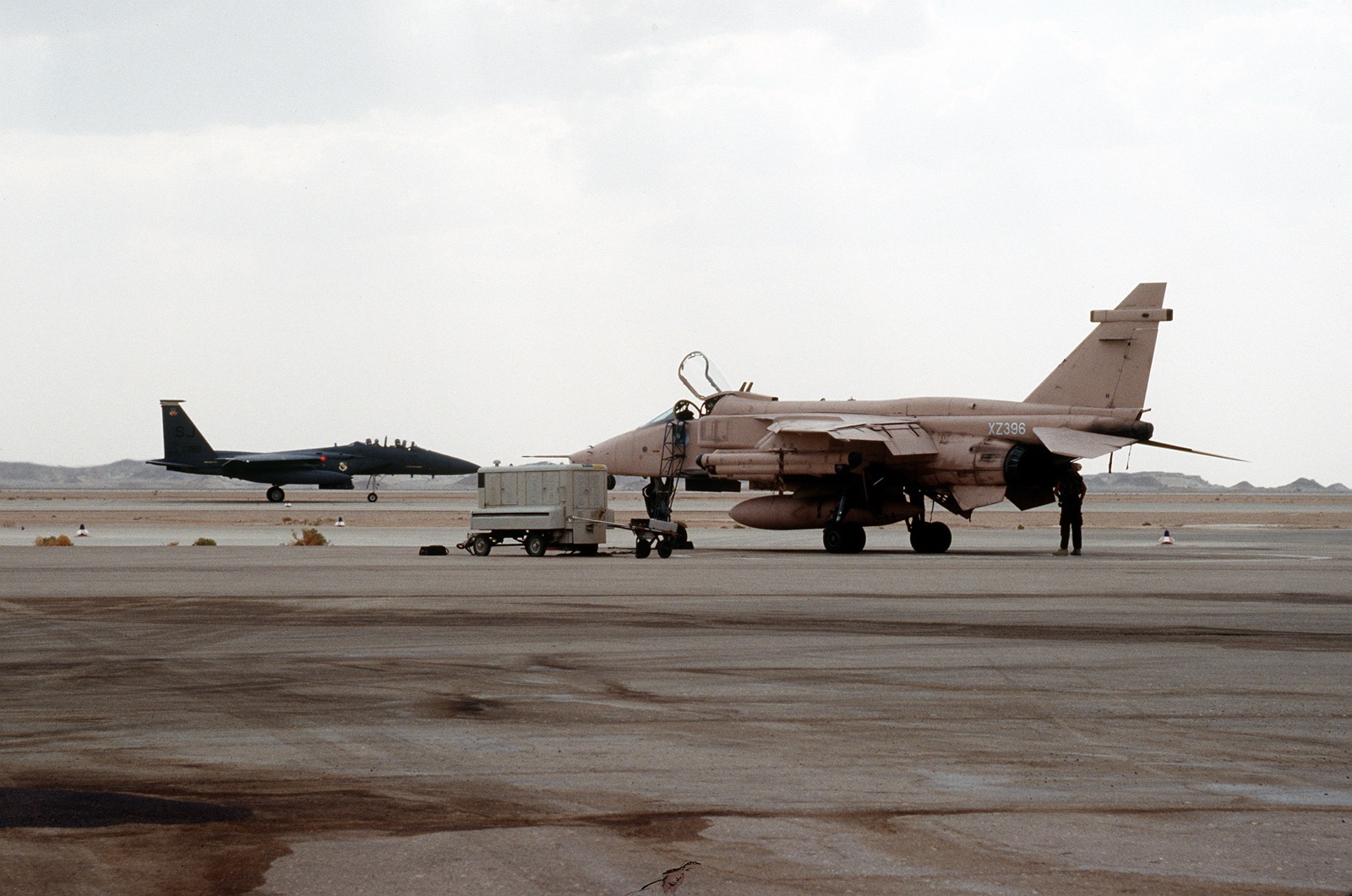 A desert-pink RAF Jaguar, XZ396, being serviced on a flight line while a dark grey F-15E taxis behind