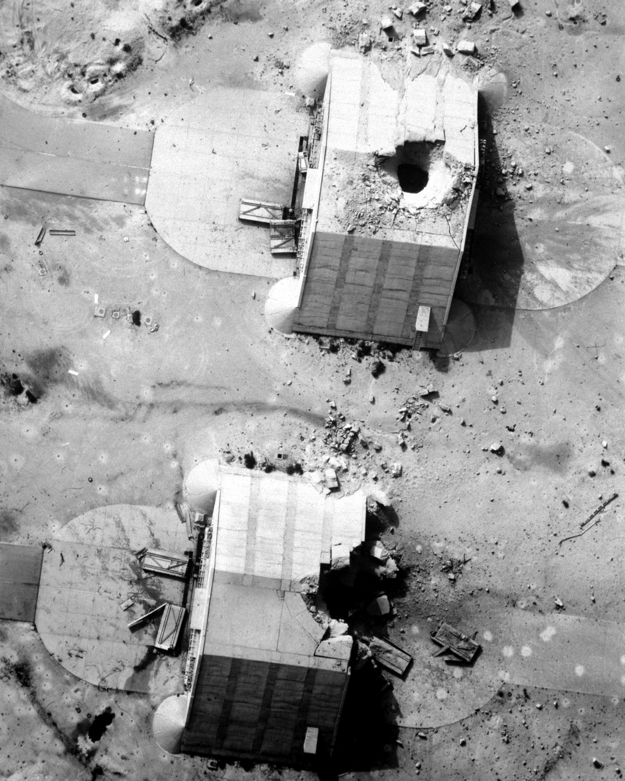 Overhead reconnaissance photograph of two concrete aircraft shelters, one with a neat hole punched through its roof, the other with its rear wall blown out