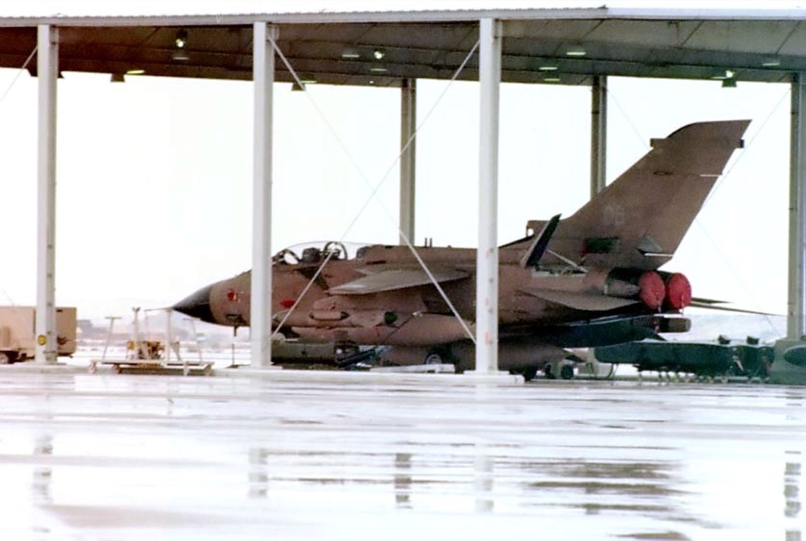A desert-pink RAF Tornado GR1, wings swept back, parked under an open-sided sunshade on a wet apron