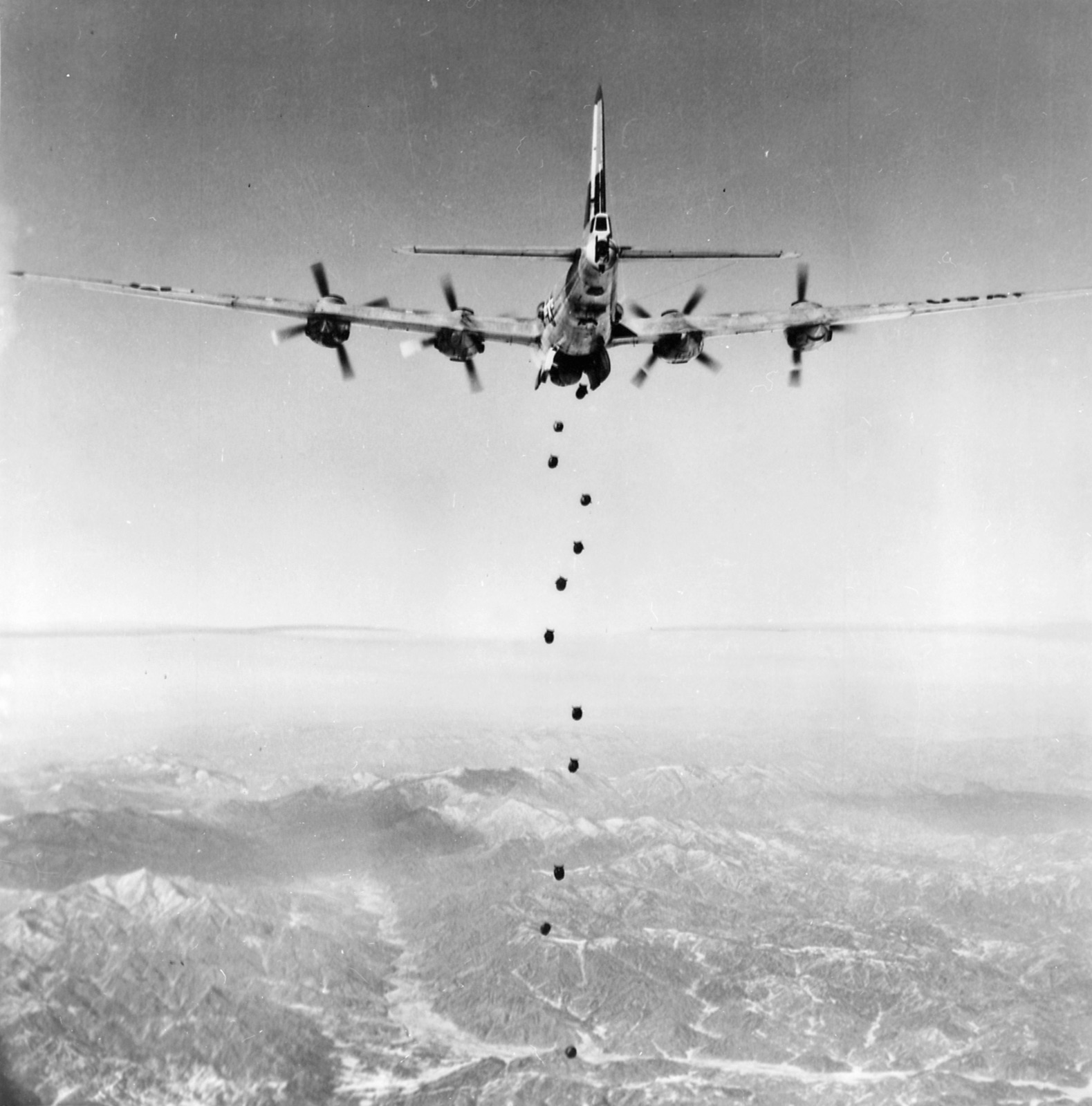 A B-29 Superfortress seen from behind and above, a vertical stick of bombs falling from its bomb bay towards snow-streaked mountains.