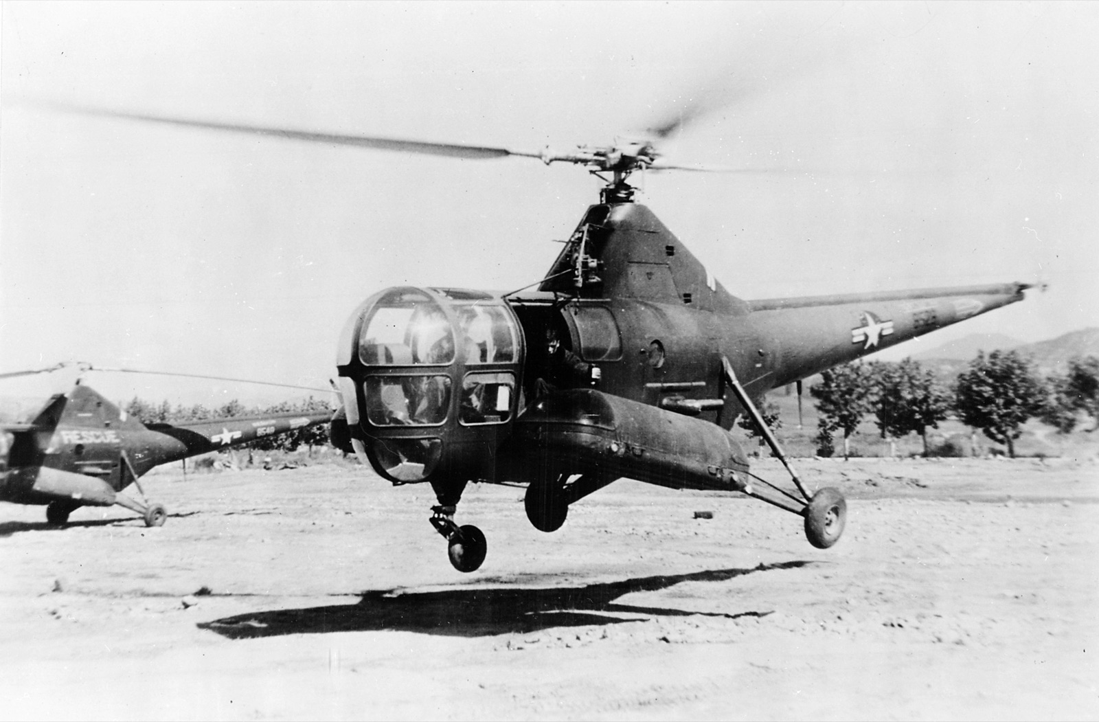 A Sikorsky H-5 helicopter lifting off a dirt field in Korea, with a second H-5 marked RESCUE parked on the left.