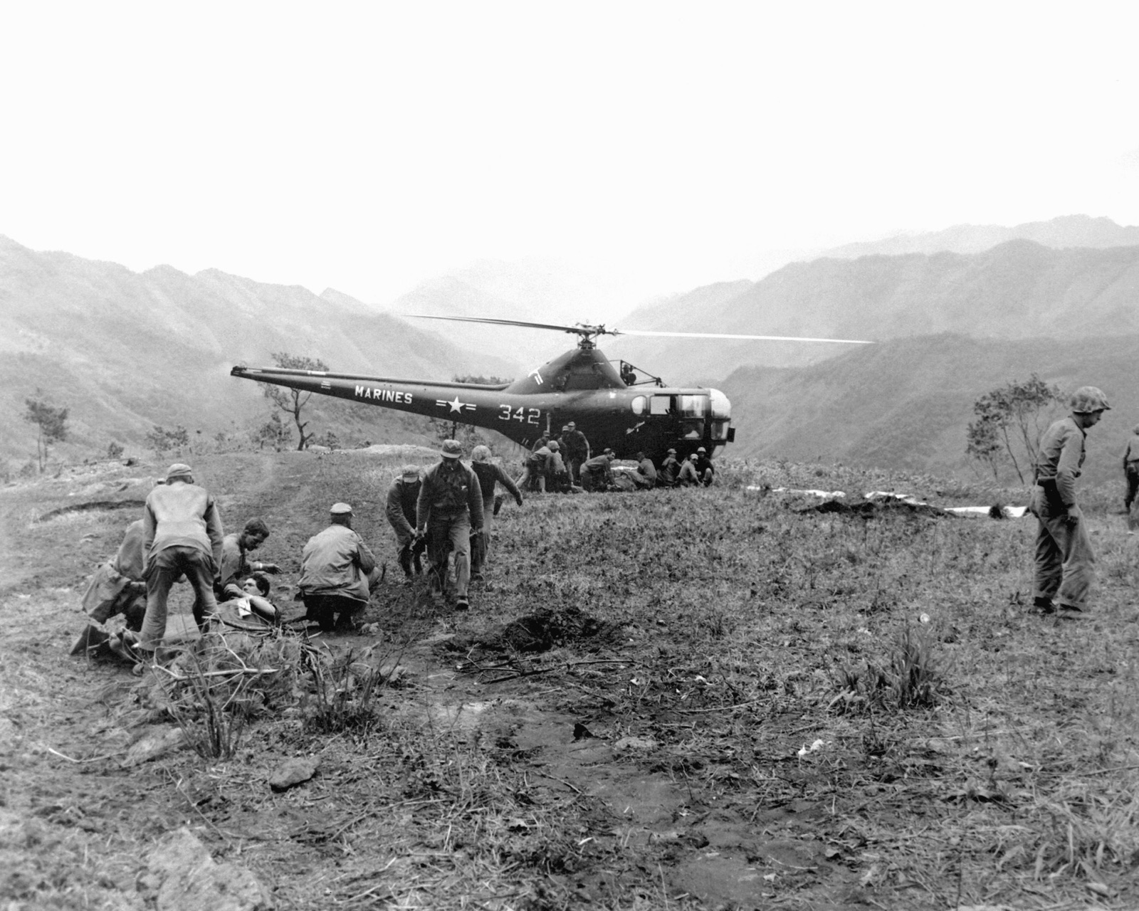 A US Marine Corps HO3S helicopter, number 342, on a hillside landing zone in Korea while Marines and Navy corpsmen tend wounded men in the foreground.