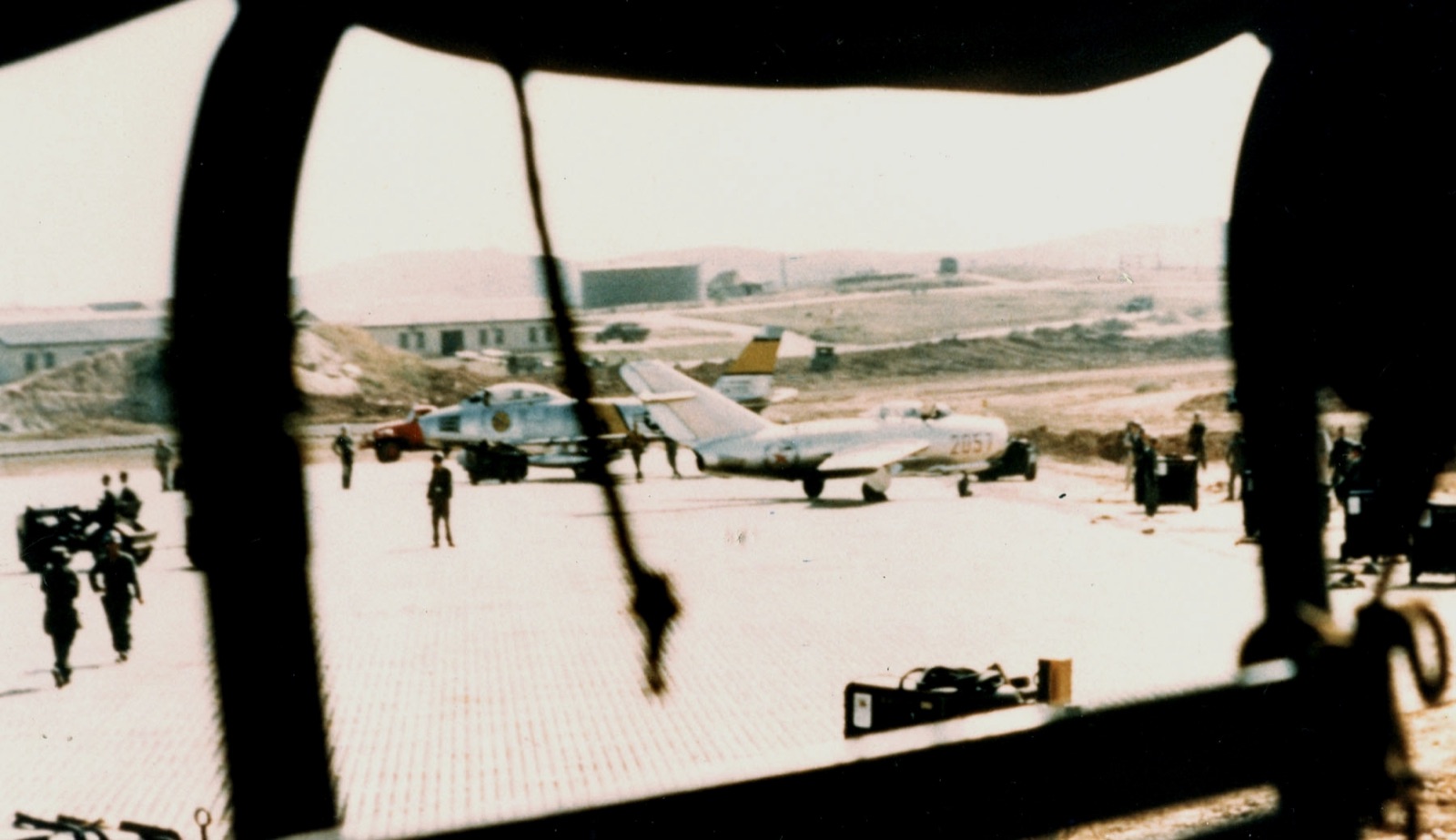 A silver MiG-15 with red star markings and the number 2057, parked on the steel-plank apron at Kimpo, photographed through a dark window frame, with a Sabre and ground crew nearby.