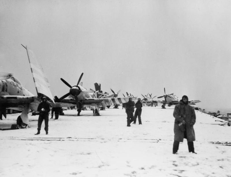 Black-and-white photograph of the snow-covered flight deck of HMS Theseus with Sea Fury and Firefly aircraft parked in rows and sailors in duffel coats.