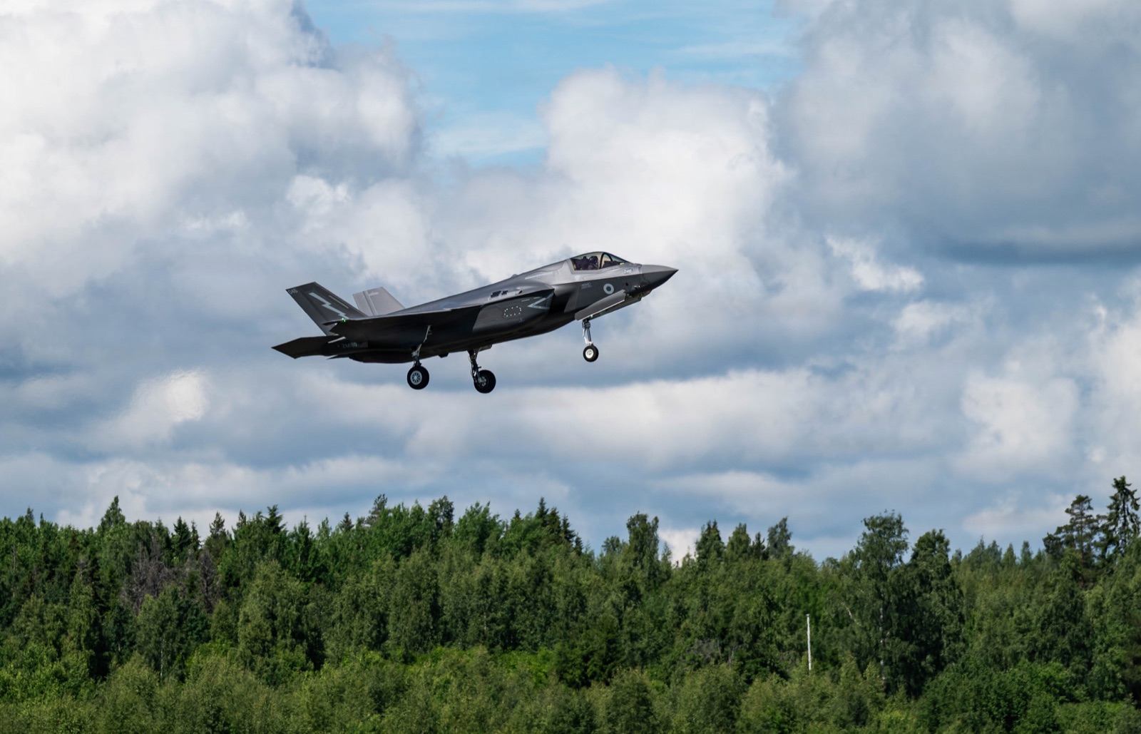 A dark grey F-35B with a lightning-bolt fin badge, gear down, flying low over a pine forest