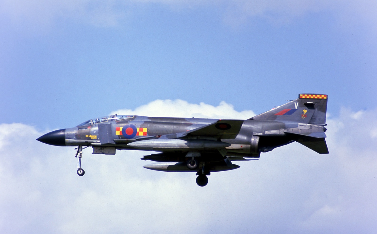 A grey-green camouflaged RAF Phantom with red and yellow squadron chequers, wheels down on approach