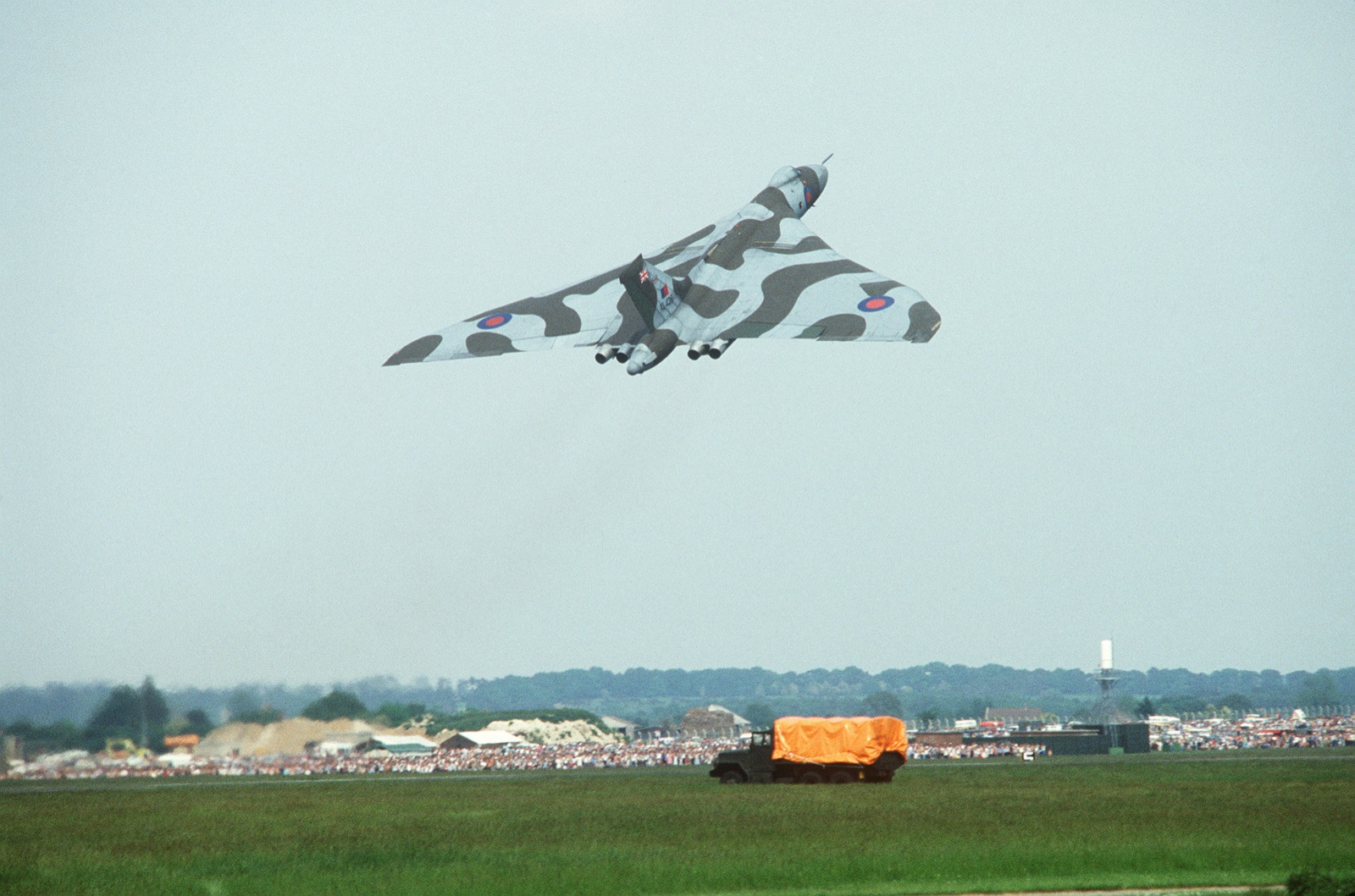 A camouflaged Avro Vulcan delta bomber climbing steeply away over a crowded airfield