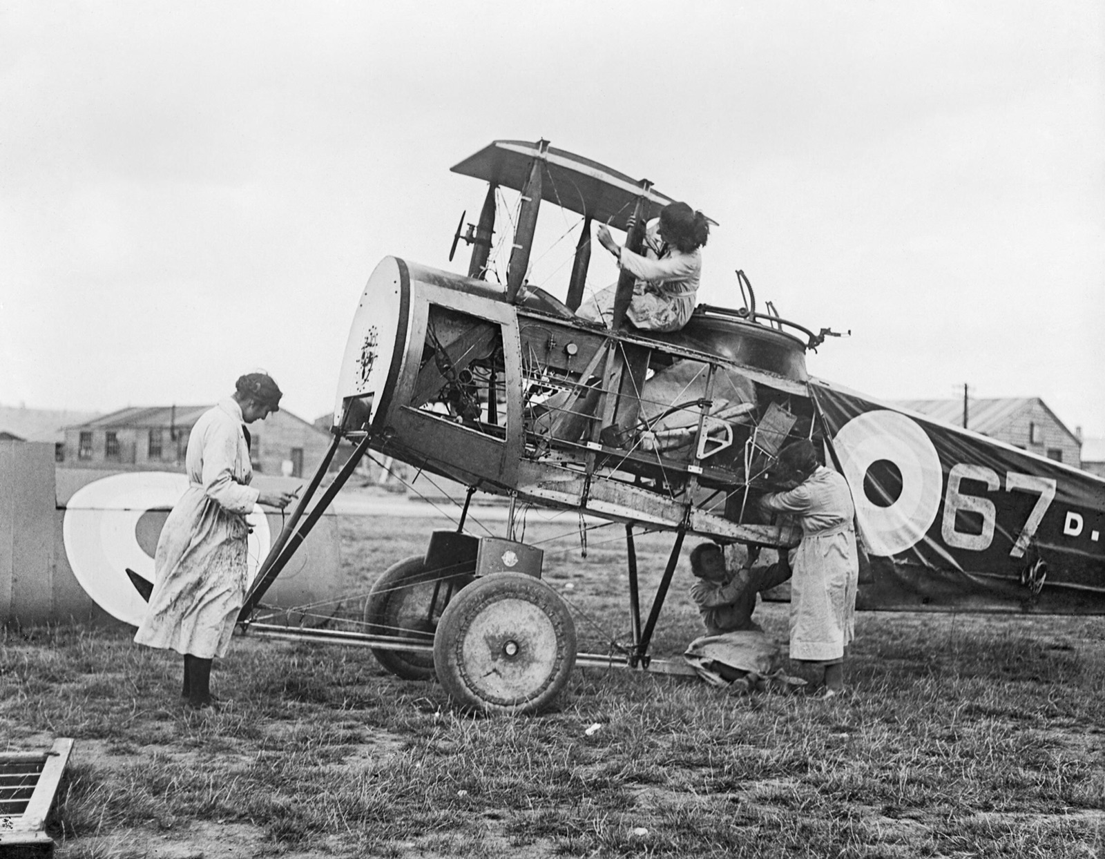 Three women air mechanics in long overalls work on a stripped-down biplane fuselage, a wing with a roundel propped beside it