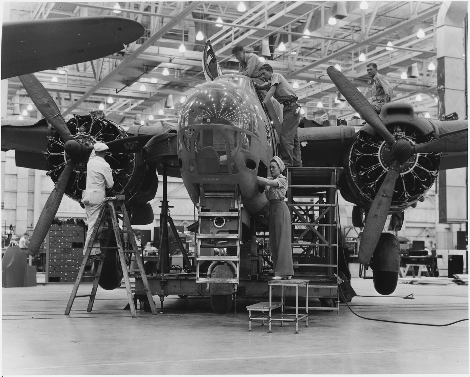 A Douglas A-20 attack bomber on the assembly line at Long Beach, men and a woman worker on ladders and steps around its glazed nose and two radial engines