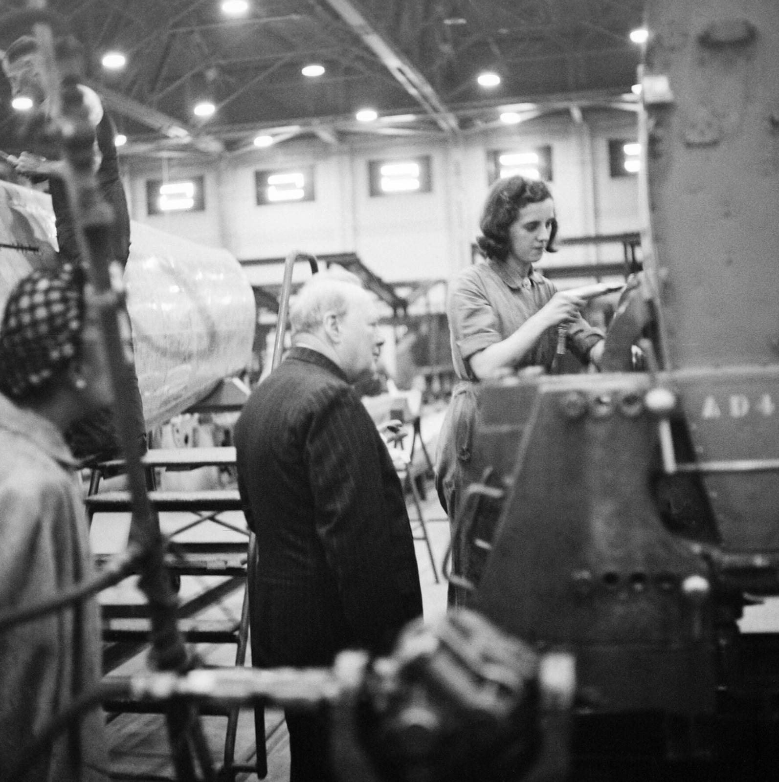 Winston Churchill, in a pinstripe suit, watches a young woman in overalls riveting on a Spitfire assembly jig in a large factory hall