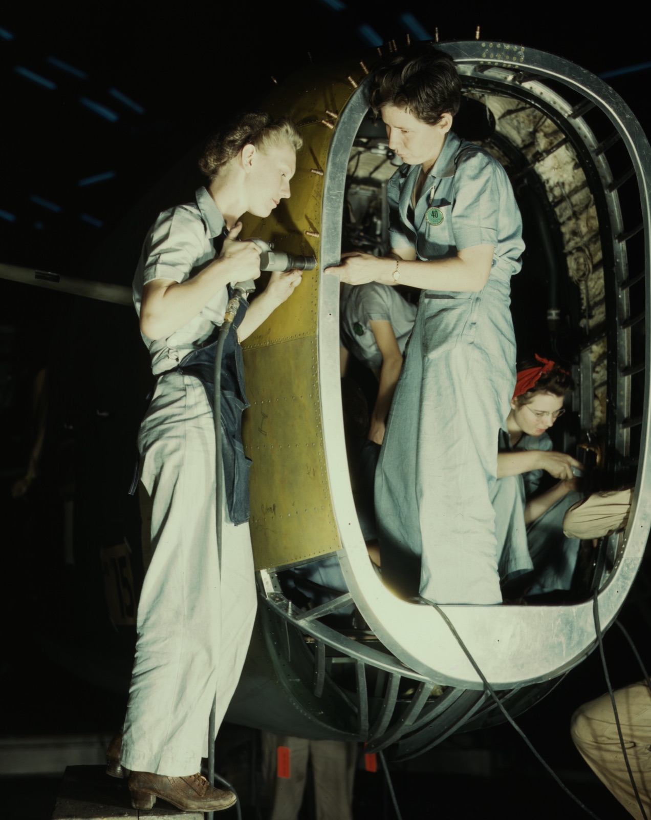 Colour photograph of two women in overalls riveting the rear fuselage section of a B-24 Liberator, a third woman in a red headscarf working inside