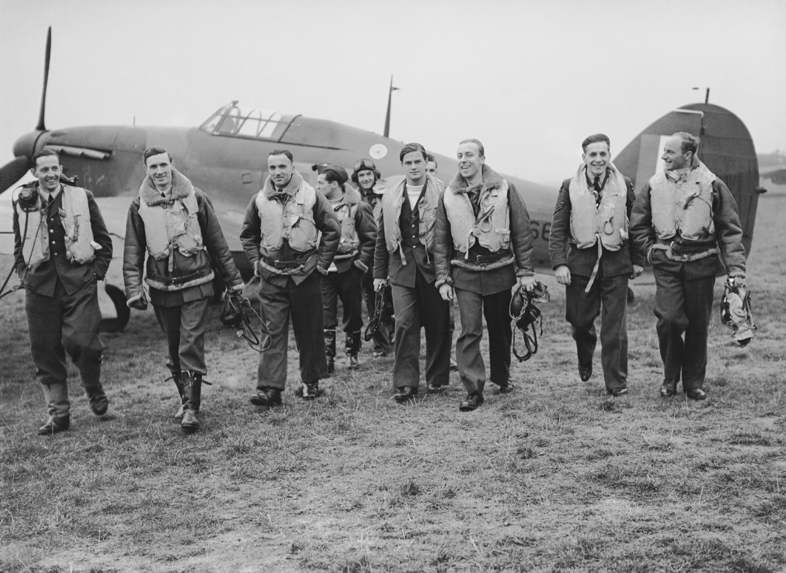 Nine pilots in Mae West life jackets and flying boots walk towards the camera, smiling, in front of a parked Hawker Hurricane
