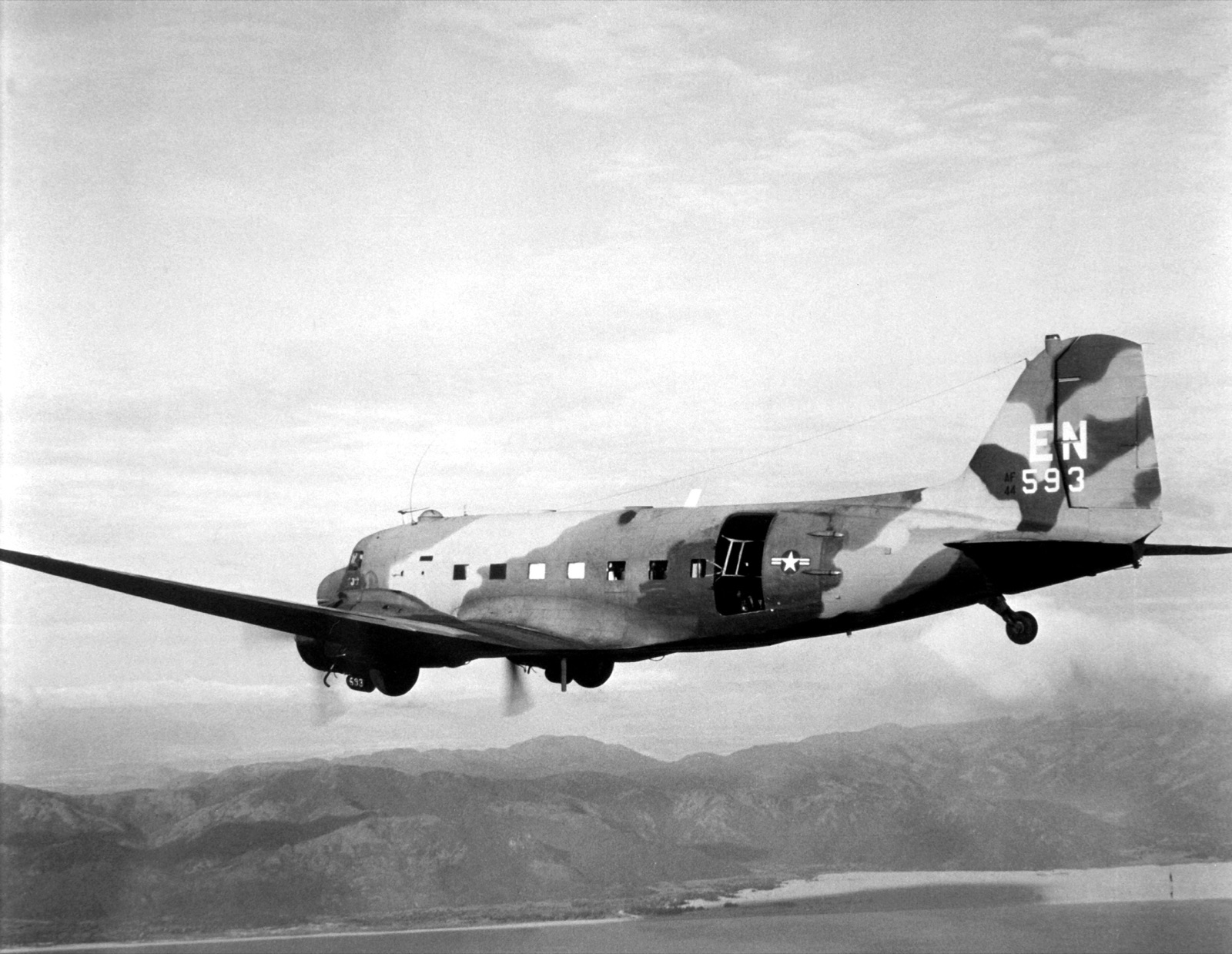 A camouflaged Douglas AC-47D gunship, tail code EN and number 593, flying over a coastline with mountains behind.