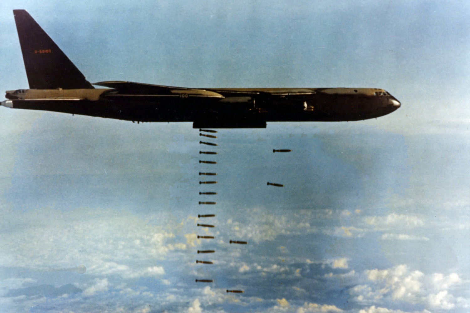A B-52D in black and camouflage paint releasing a long vertical stream of bombs over cloud.
