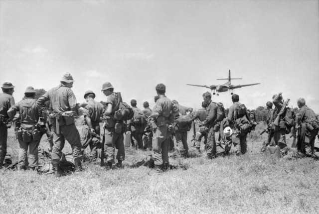 Australian infantrymen with packs stand on a grass strip watching a twin-engined Caribou come in to land.