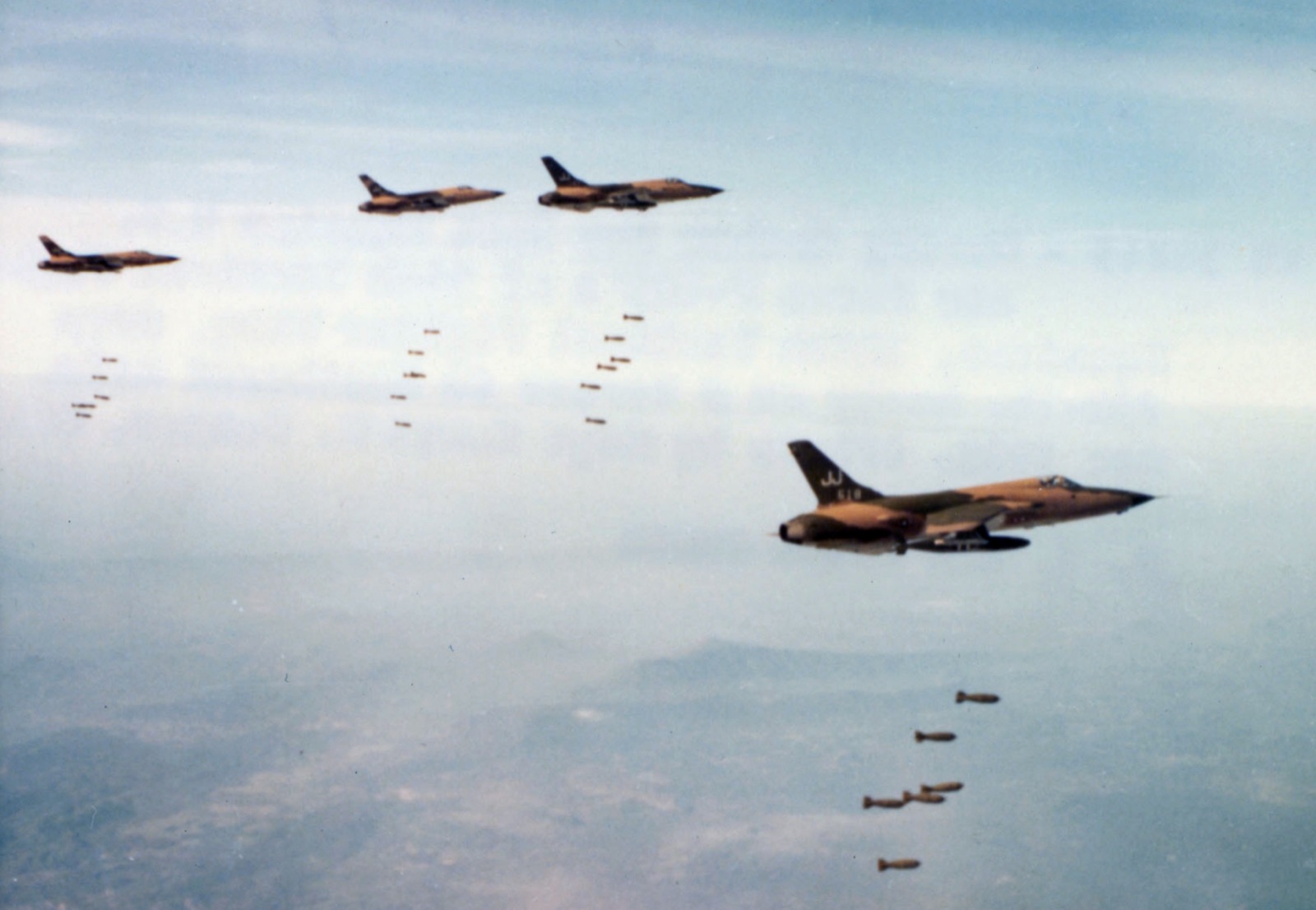 Four camouflaged F-105D Thunderchiefs releasing bombs in formation above hazy countryside.