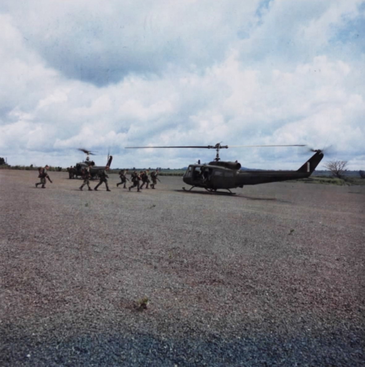 Australian soldiers with packs and rifles run across a gravel pad towards two olive-drab RAAF Iroquois helicopters.