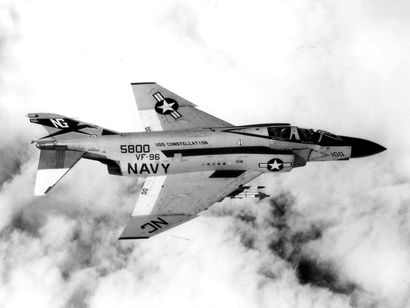 Overhead view of a US Navy F-4J Phantom of VF-96, number 100 and serial 5800, marked USS Constellation, flying above broken cloud with missiles on an underwing pylon.