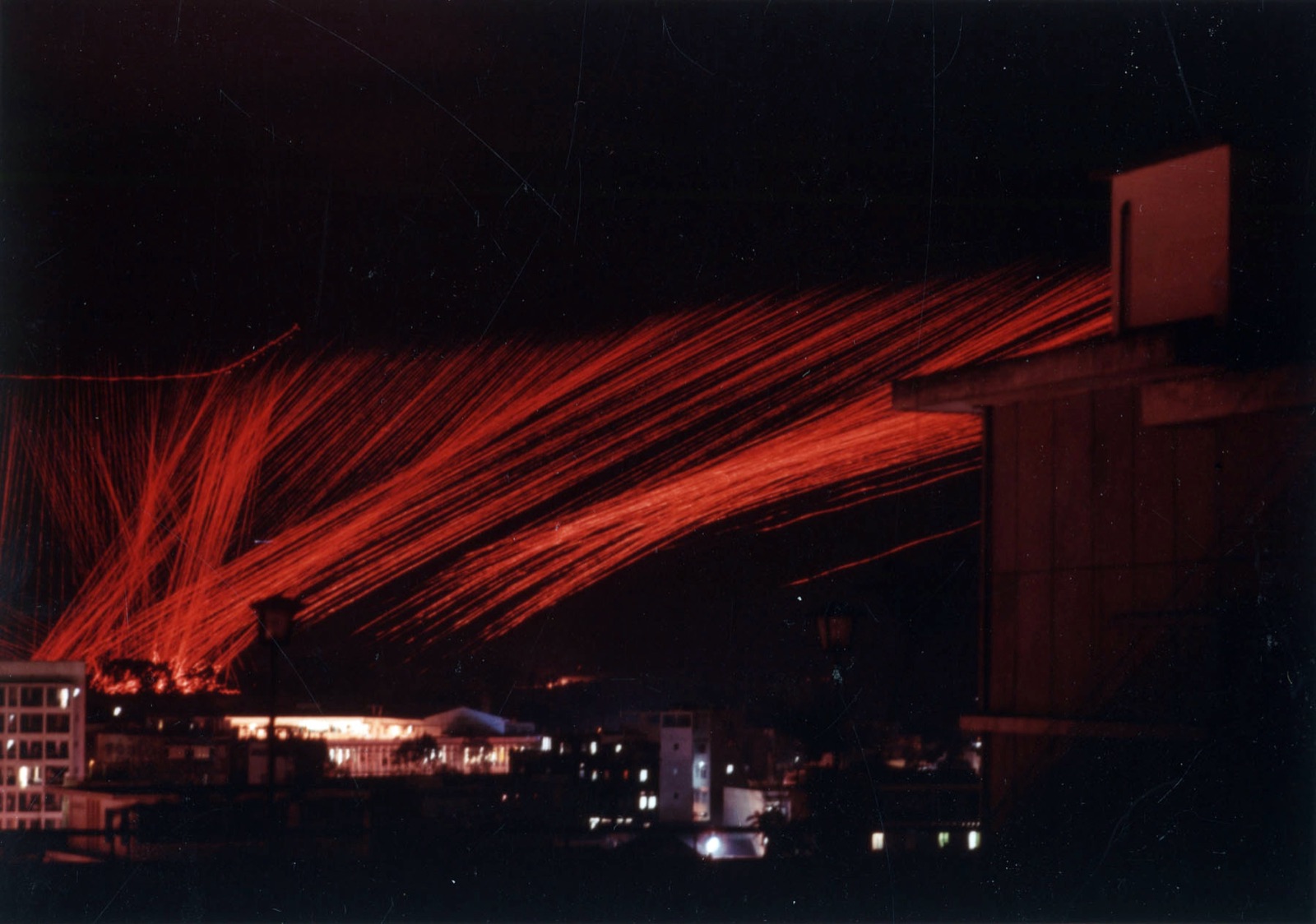 Time-exposure photograph at night of curving red tracer streams from an AC-47 gunship above the lit buildings of Saigon.