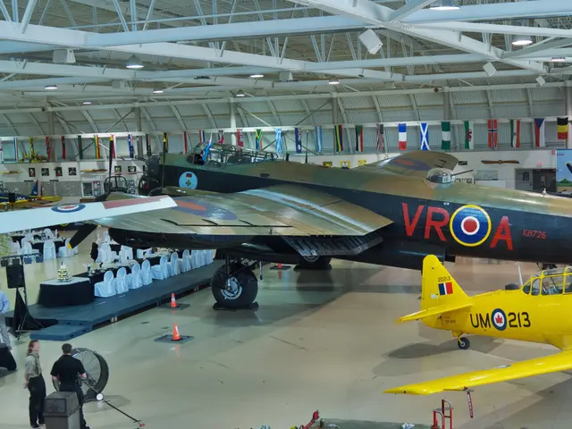 Dark green and brown Avro Lancaster bomber with red VR-A codes parked inside a bright hangar, with a yellow training aircraft in front