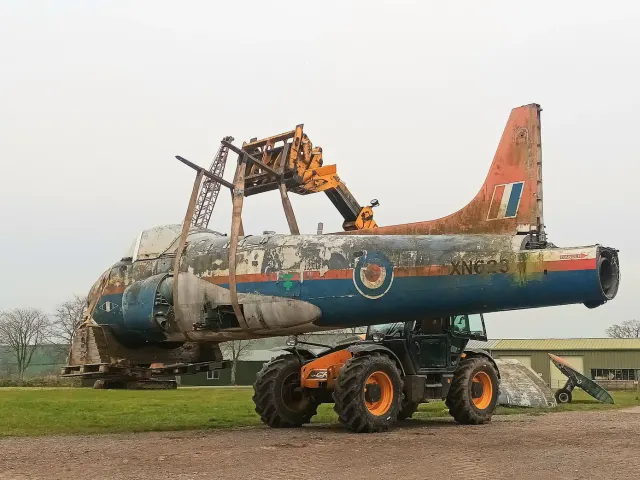 Weathered blue, grey and orange Jet Provost fuselage lifted on a telehandler beside a farm building