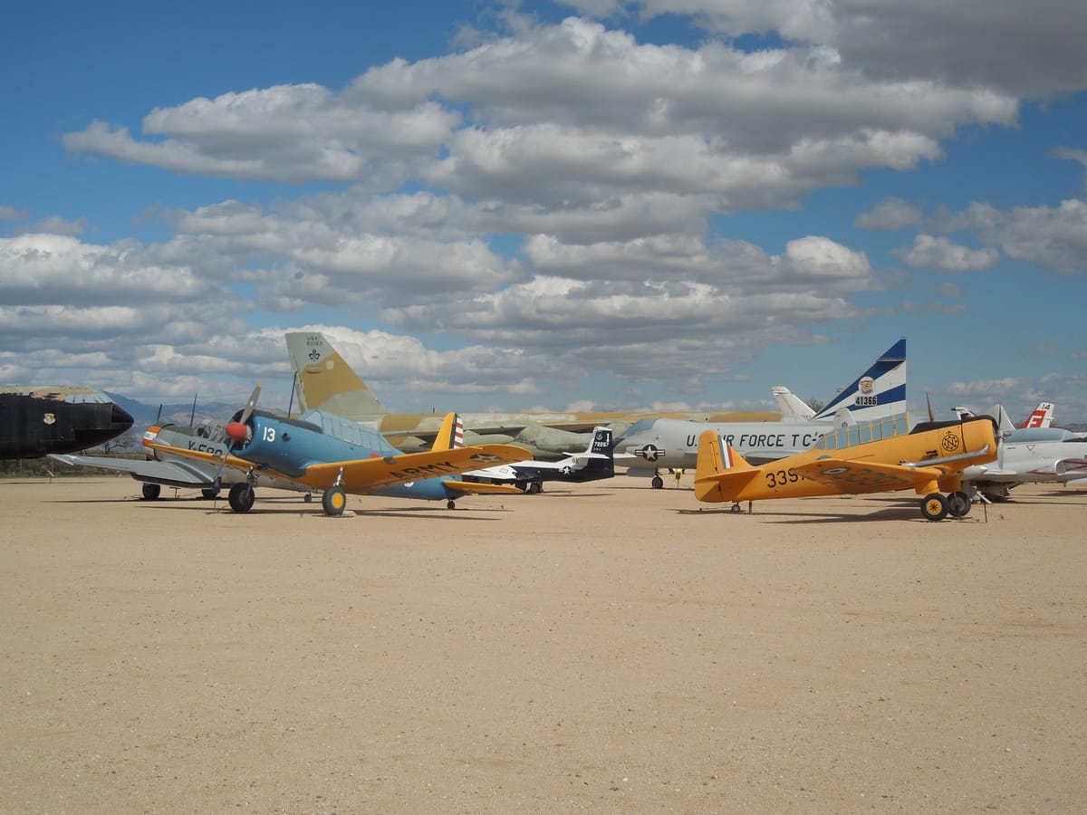 Lines of propeller aircraft parked on sandy ground beneath scattered white clouds