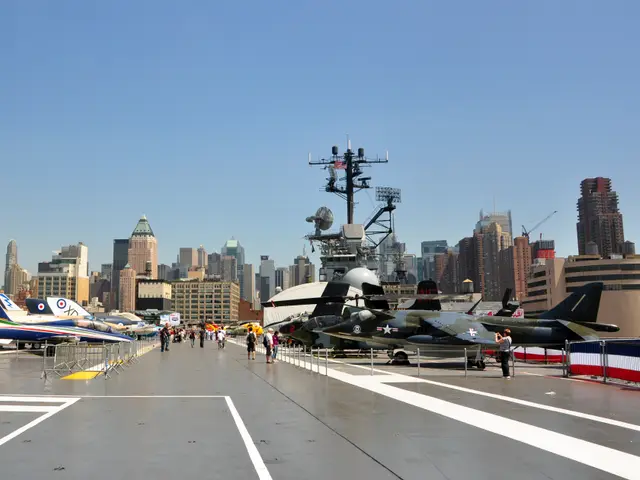 View along the carrier's flight deck with rows of jets, the island and Midtown skyscrapers under a blue sky